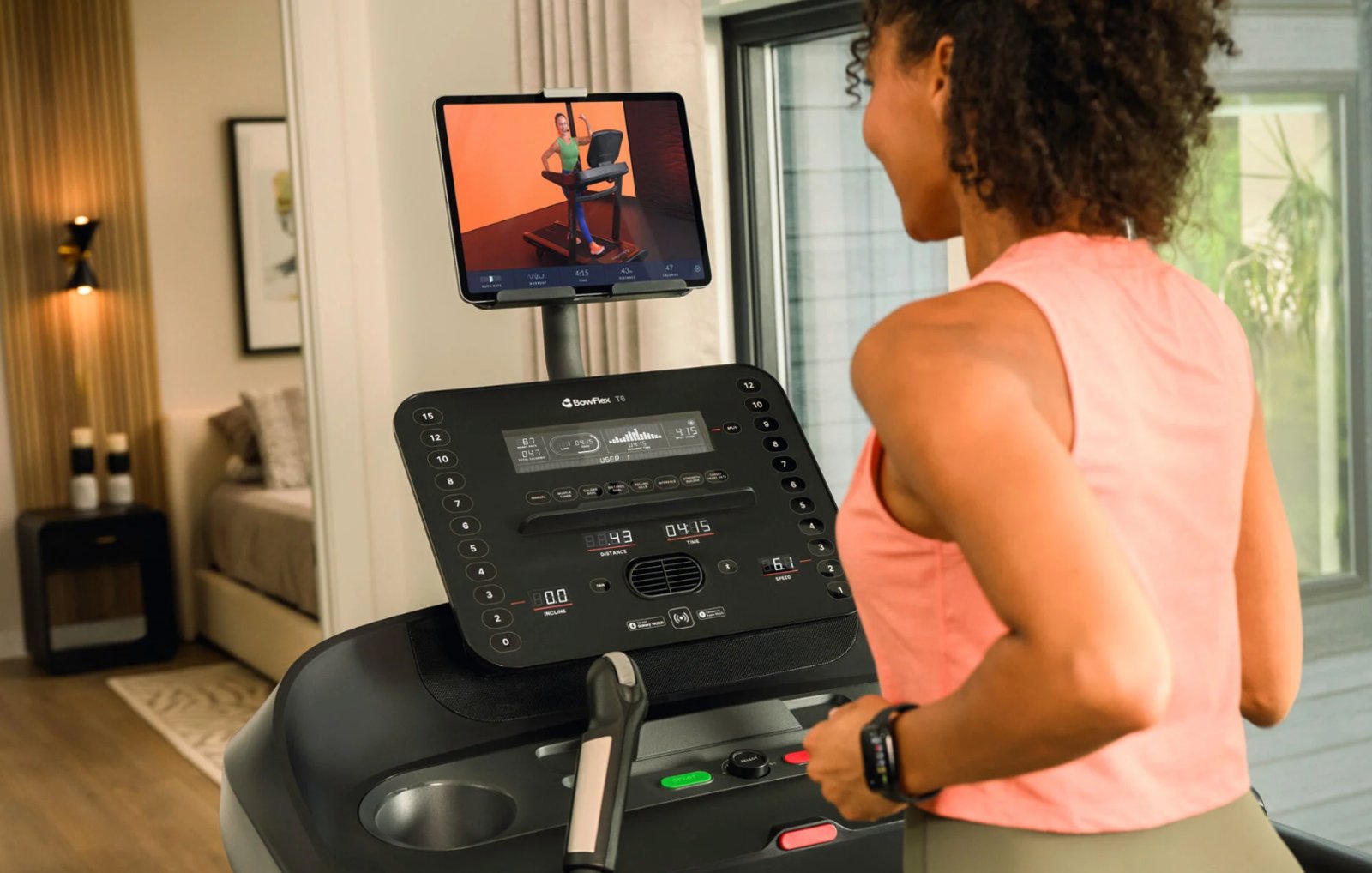 A runner on the BowFlex T6 treadmill in a bright living room, following a trainer-led workout on a tablet mounted above the console, with the LED display showing distance, time, speed, incline, heart rate and calories.