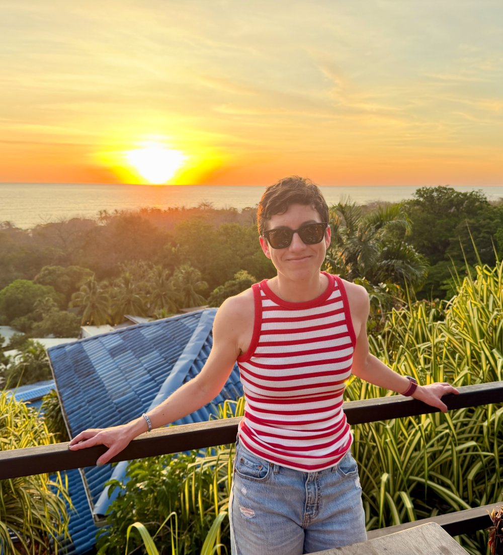 Edyta Blaszczyk smiling at sunset, wearing a red and white striped tank top, with the ocean and tropical greenery behind her.
